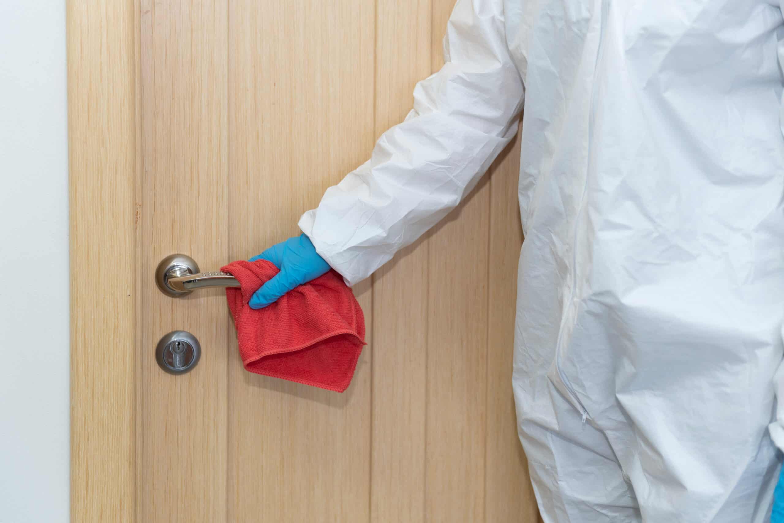 A woman in protective gloves and suit holding a red cloth cleaning the door knob